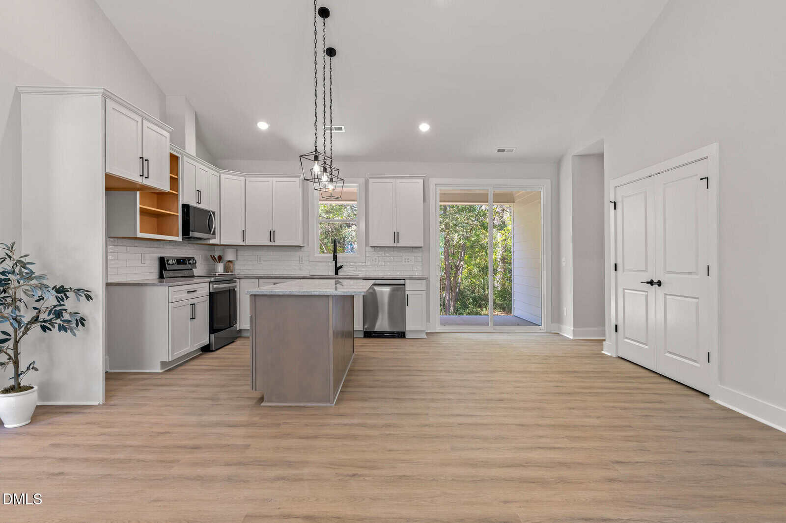Modern kitchen with white cabinets, an island, stainless steel appliances, pendant lighting, light wood flooring, large double doors, and sliding glass doors opening to a green outdoor area. Potted plant in the corner.