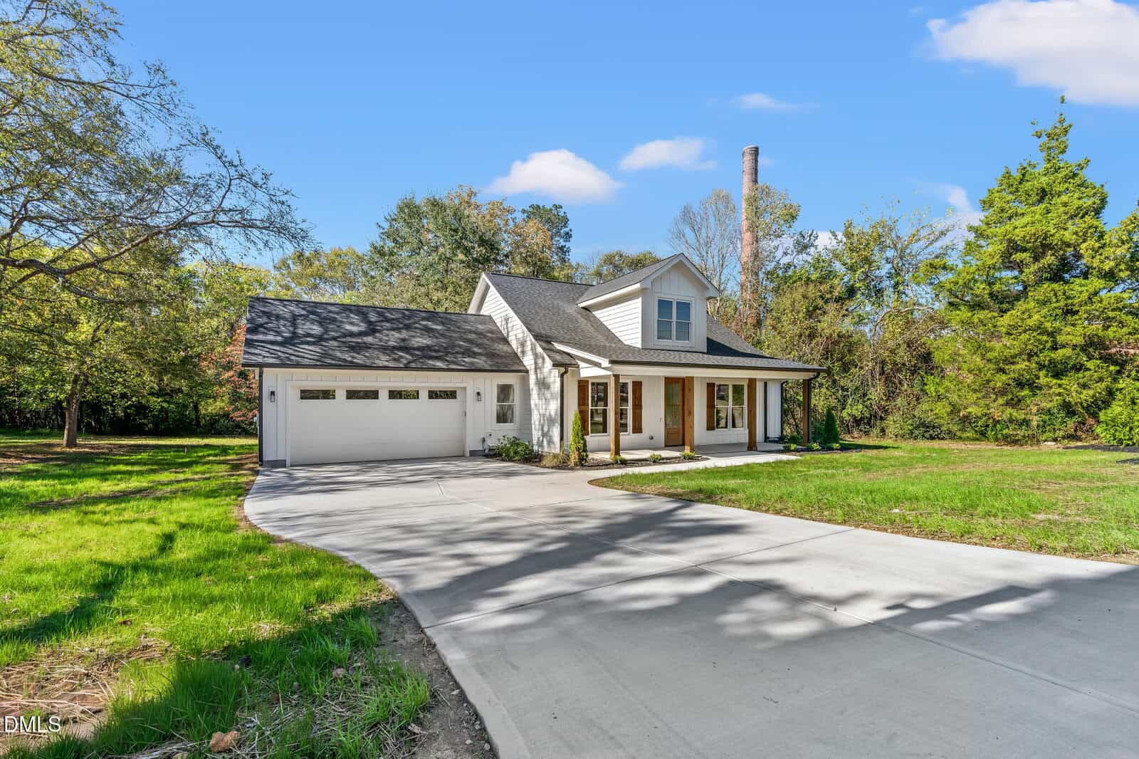A white, two-story house with a front porch and attached two-car garage sits next to a large driveway, surrounded by green grass, trees, and a clear blue sky.