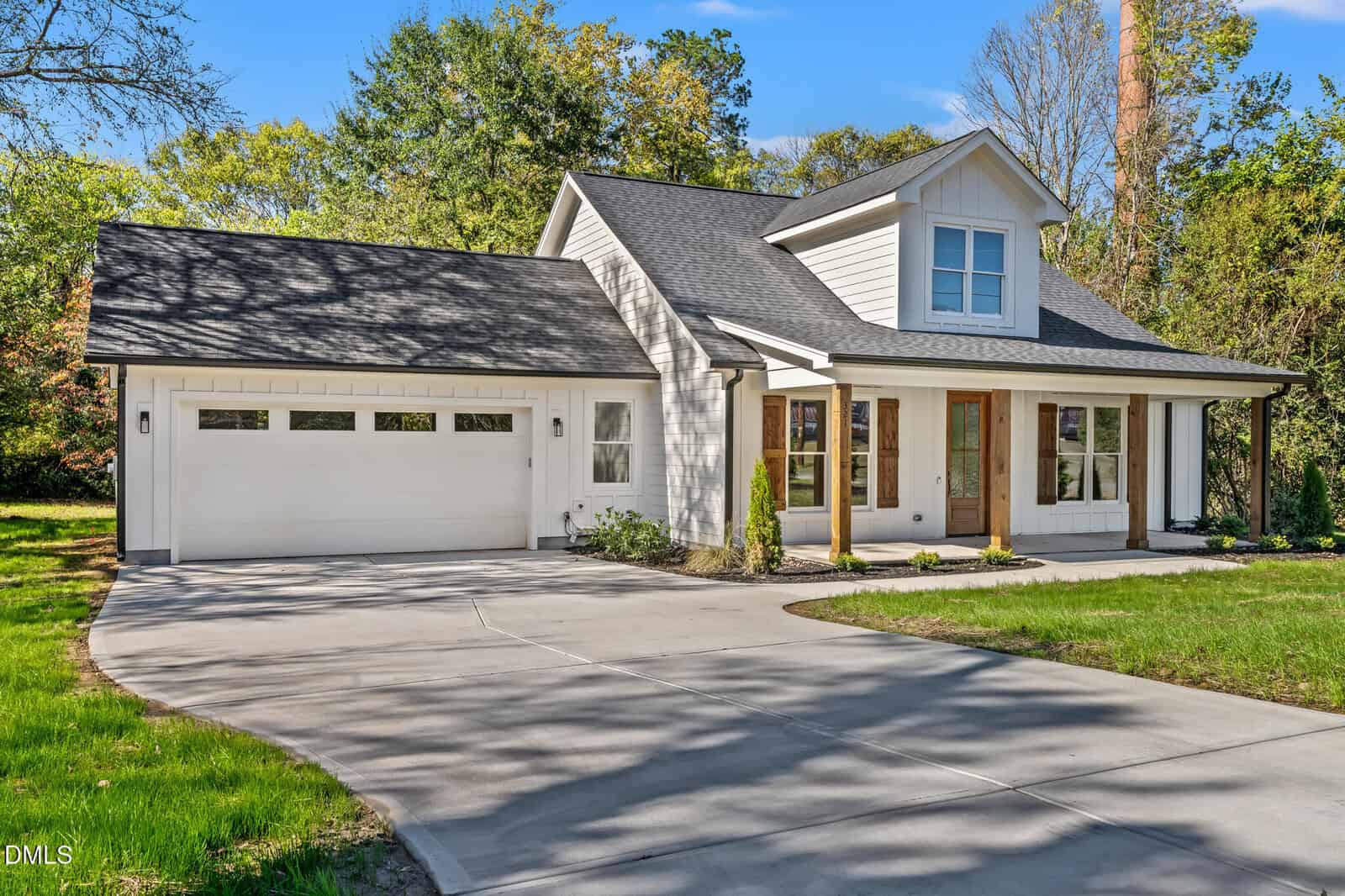 Modern white house with black roof, attached two-car garage, large front windows, and wooden pillars on a covered porch, surrounded by green lawn and trees under a clear blue sky.