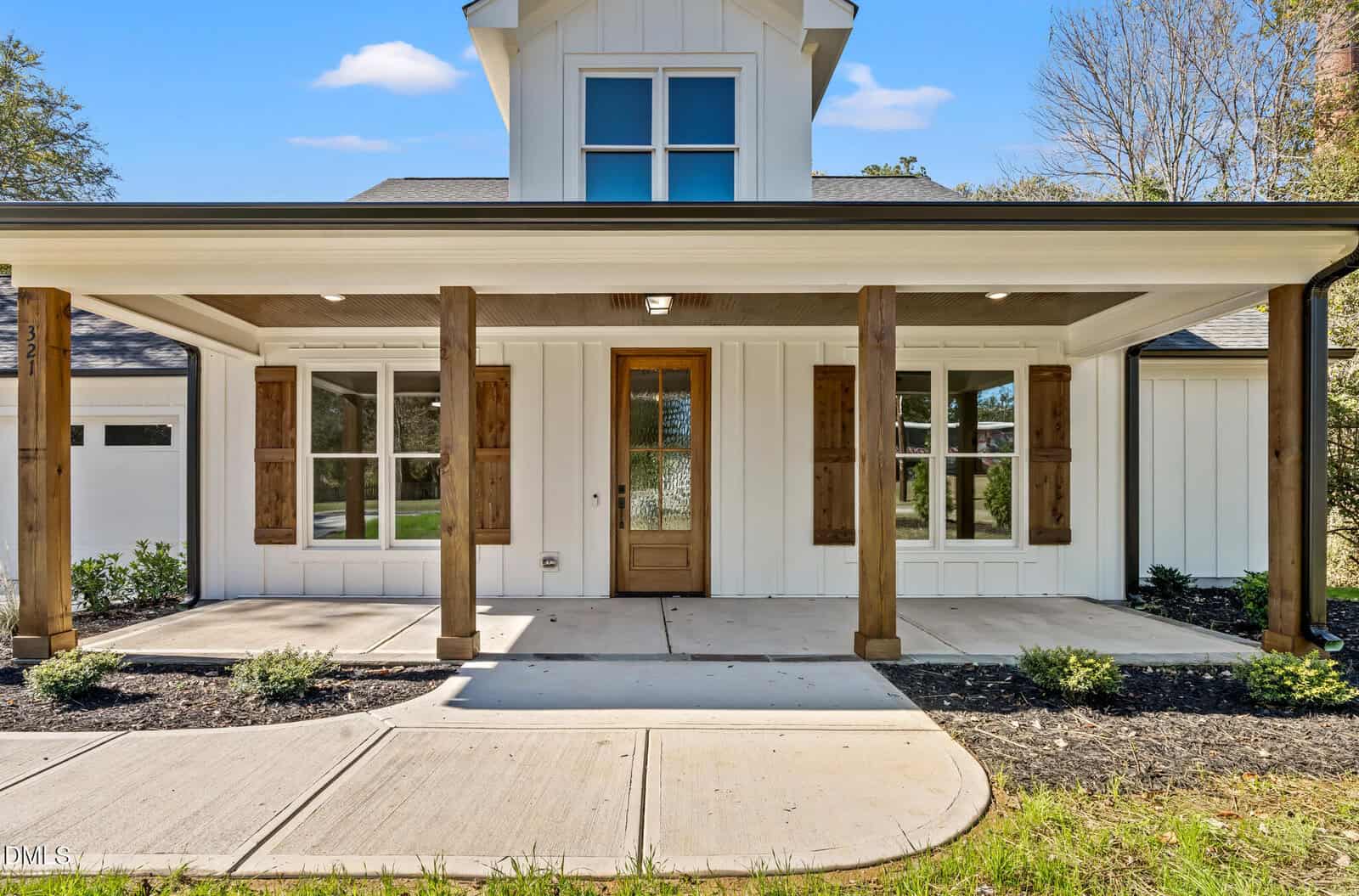 A modern white house with wooden columns, a covered front porch, wooden shutters, a glass-paneled front door, and landscaped flower beds along a curved concrete walkway.