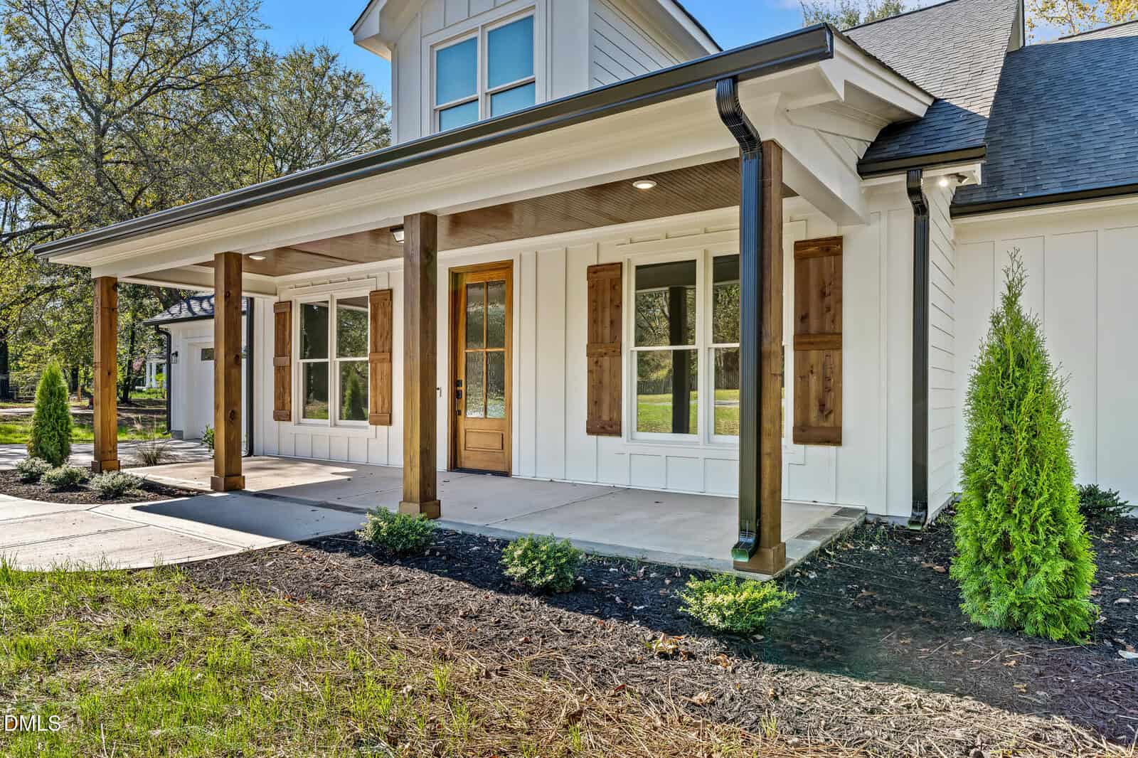 A modern farmhouse-style home with white siding, a wooden front door, wood shutter accents on windows, covered front porch with wood columns, and simple landscaping.