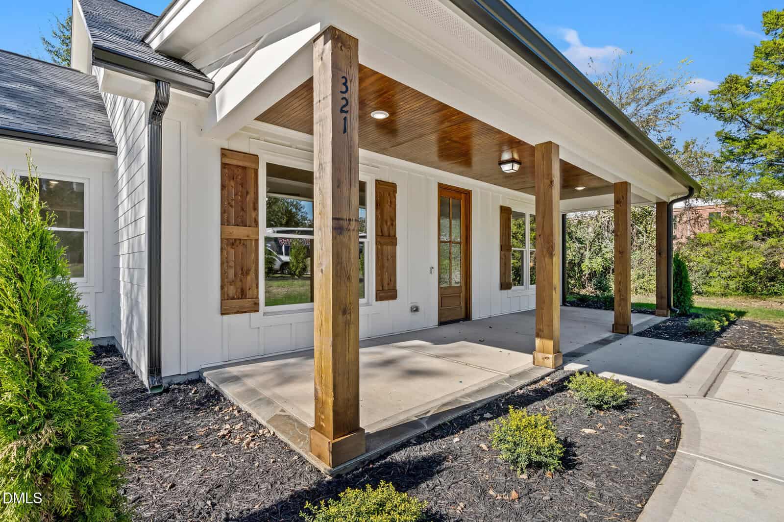 A modern farmhouse-style front porch with wooden pillars, a wooden front door, and rustic wooden shutters on white siding, surrounded by small shrubs and a concrete walkway. The house number "321" is on a pillar.