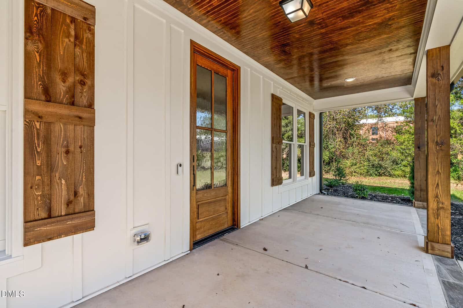 A covered front porch with a wooden door, white siding, wooden shutters, columns, and a wood-paneled ceiling; the porch overlooks a yard with trees and greenery in the background.