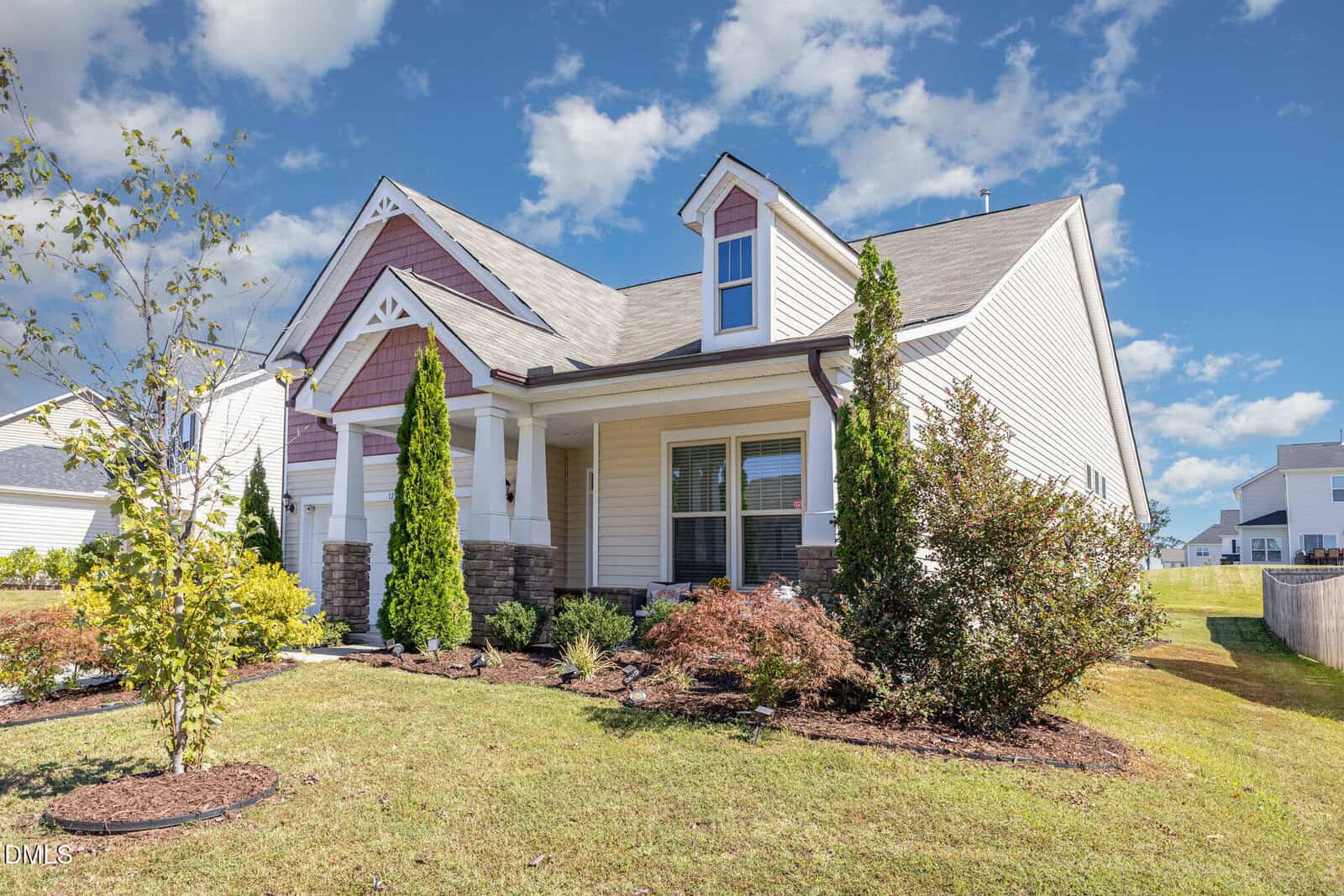 Single-story house with beige siding, white trim, and a covered front porch. There are tall shrubs, small trees, and grass in the landscaped front yard. The sky is partly cloudy and other houses are visible in the background.