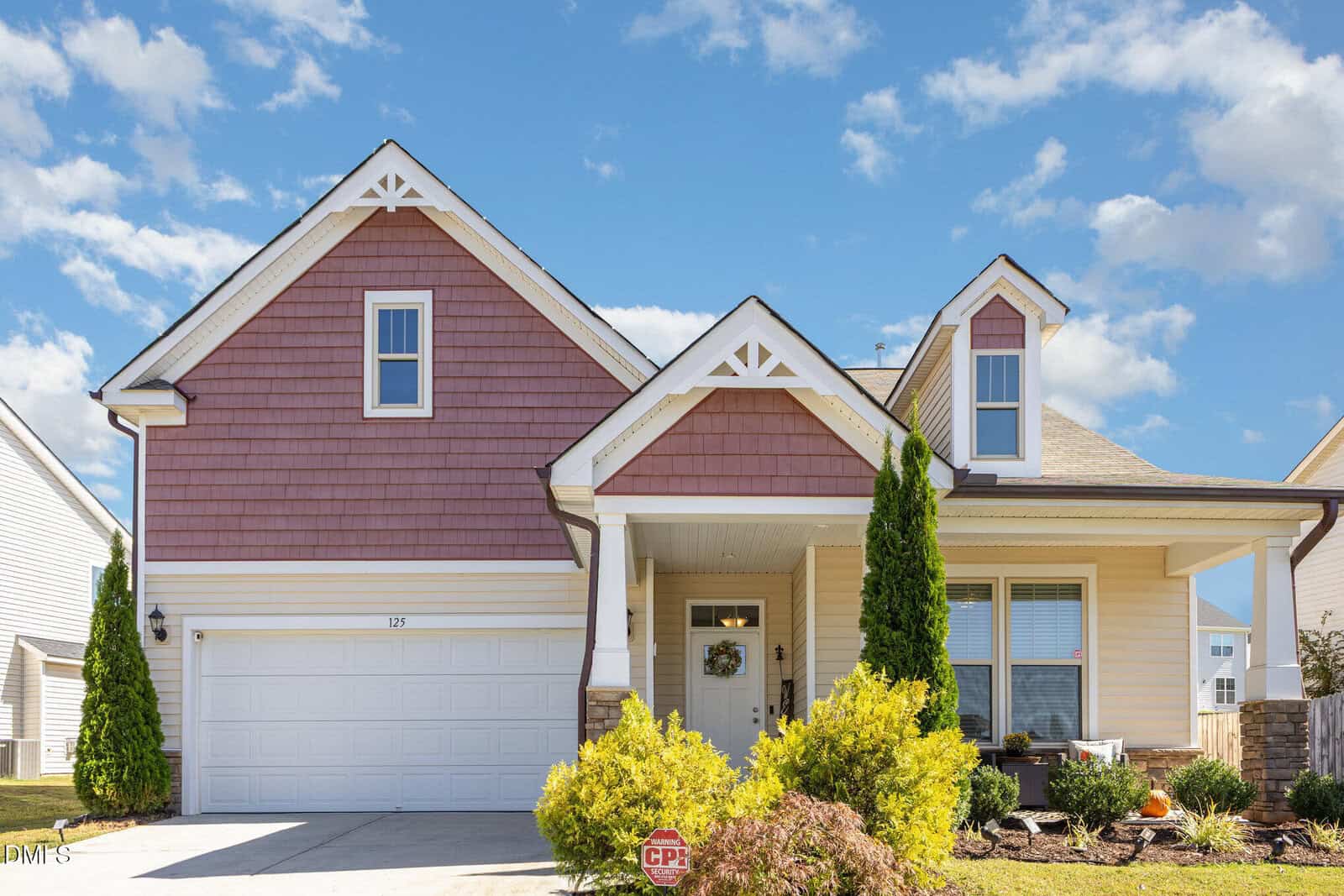 A two-story suburban house at 125 Cranes Nest Drive in Franklinton, featuring a double garage, beige siding, red-brown shingles on the upper portion, white trim, front porch, two tall shrubs, and a manicured lawn under a blue sky with scattered clouds.