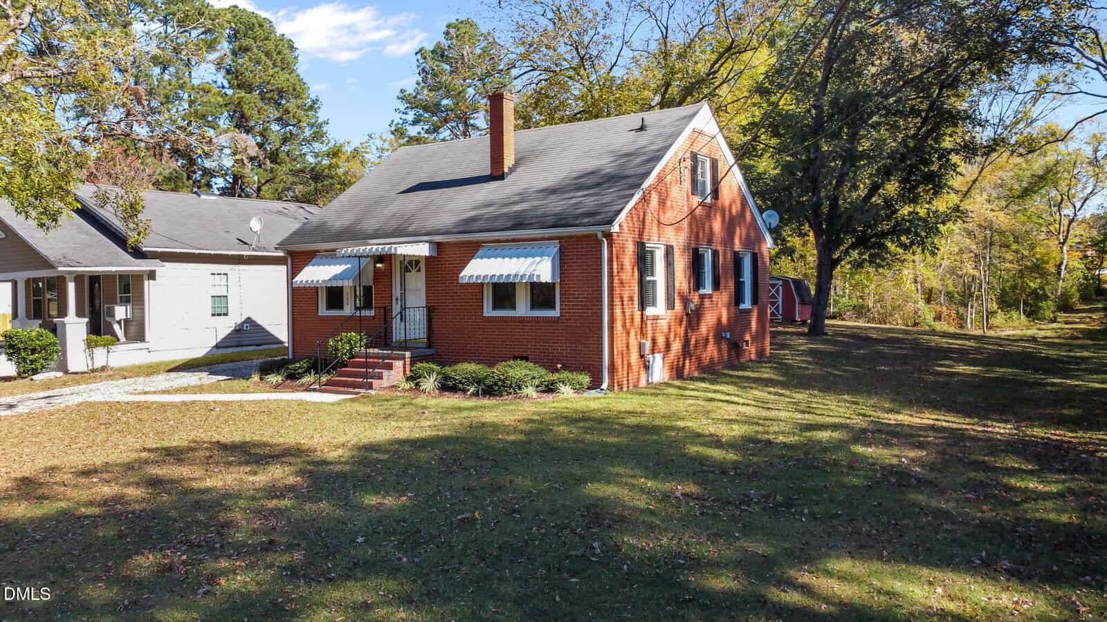 A red-brick house with a gray roof, blue-and-white awnings over the windows, a small front porch with steps, and a grassy yard. Large trees and a neighboring house are visible in the background.