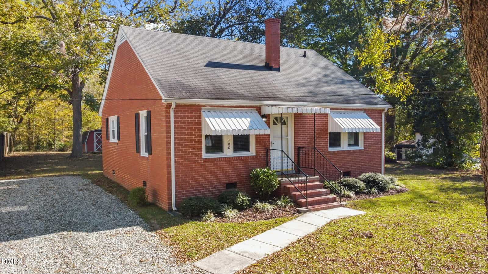A small red-brick house with a steep roof, white awnings over the windows and door, a chimney, steps leading to the entrance, and a gravel driveway on the left, surrounded by grass and trees.
