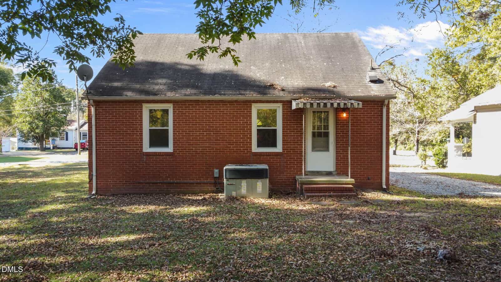 A small brick house with two windows and a door on the back side. A window air conditioning unit is installed next to the steps leading to the door. The yard is grassy with some fallen leaves and trees nearby.