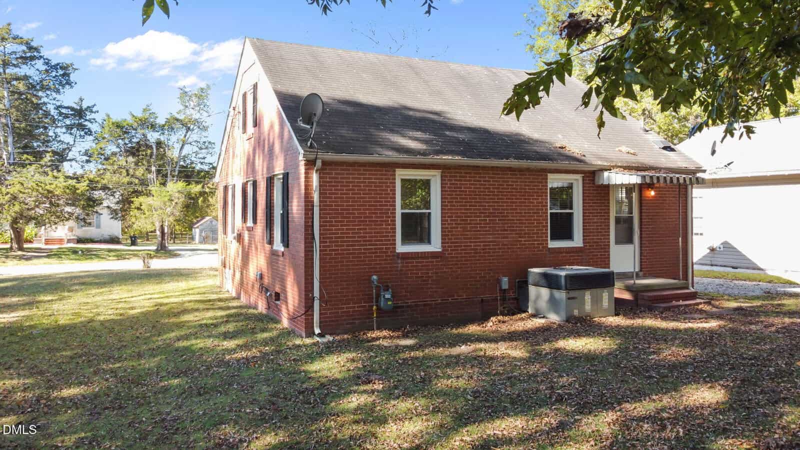 A small red-brick house with a black shingled roof, white-trimmed windows, and a back door. A large HVAC unit and a satellite dish are visible. The yard is grassy with some scattered leaves and trees nearby.