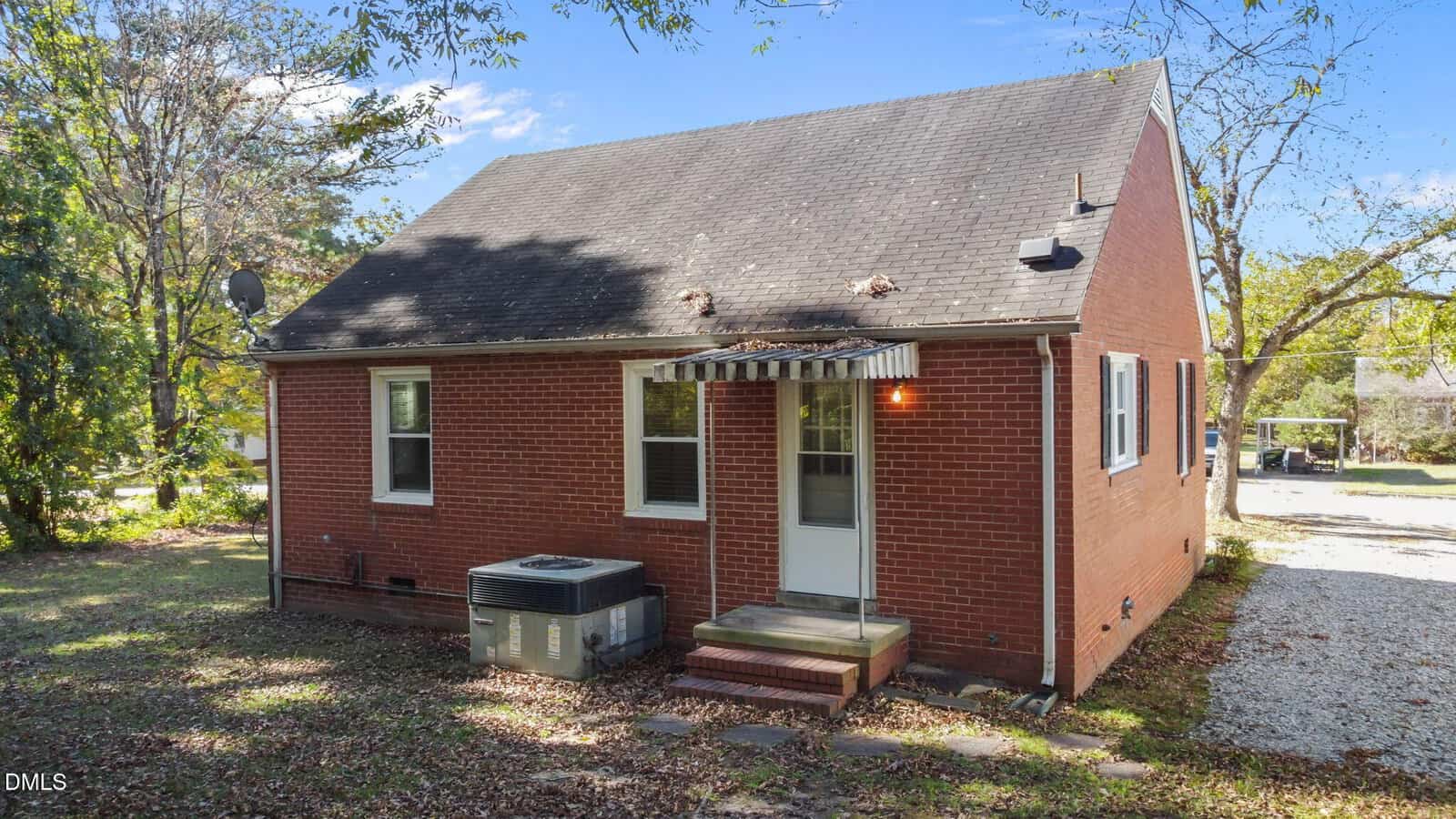 A small red brick house with a steep roof, two windows, and a white back door with a metal awning. An HVAC unit sits by the door, and there are trees, fallen leaves, and a gravel driveway visible.