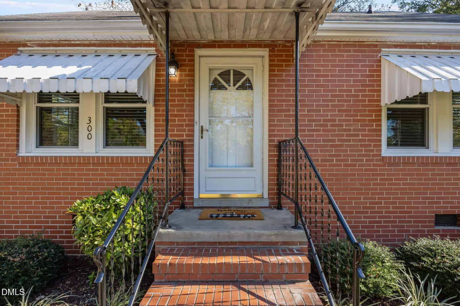A brick house with a white screen door, metal awning, and black railings leading up red brick steps. The number 300 is visible near the left window, with green shrubs on either side of the entryway.