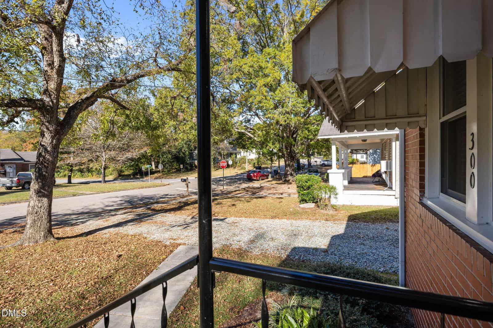 View from a porch looking out onto a suburban street with trees, parked cars, and a neighboring house. The house has a brick exterior, metal railing, and the number 300 displayed near the door.