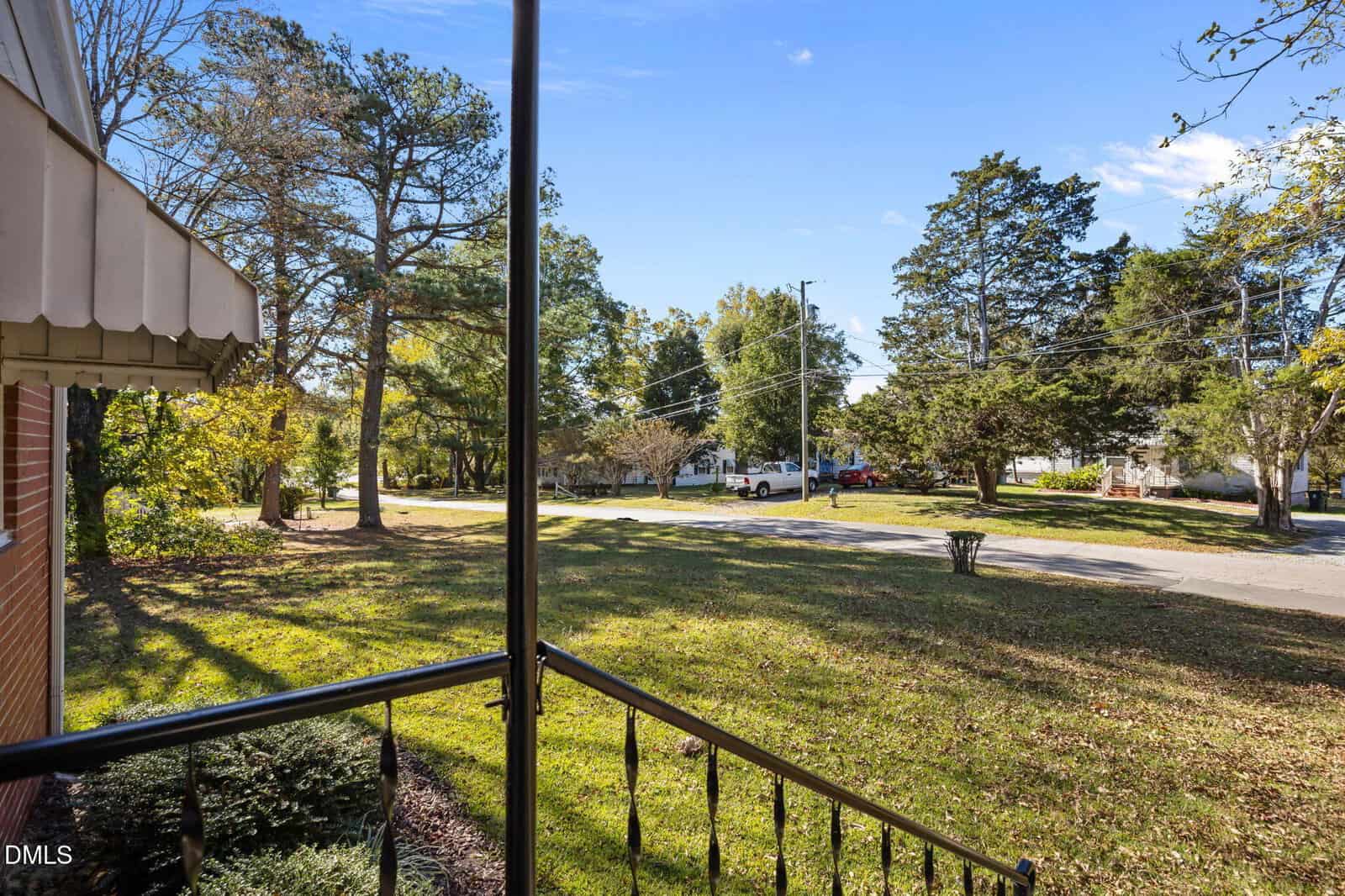 View from a porch showing a lawn with scattered trees, a street with parked cars, and houses in the background under a clear blue sky. The image includes part of a brick building and metal railing in the foreground.