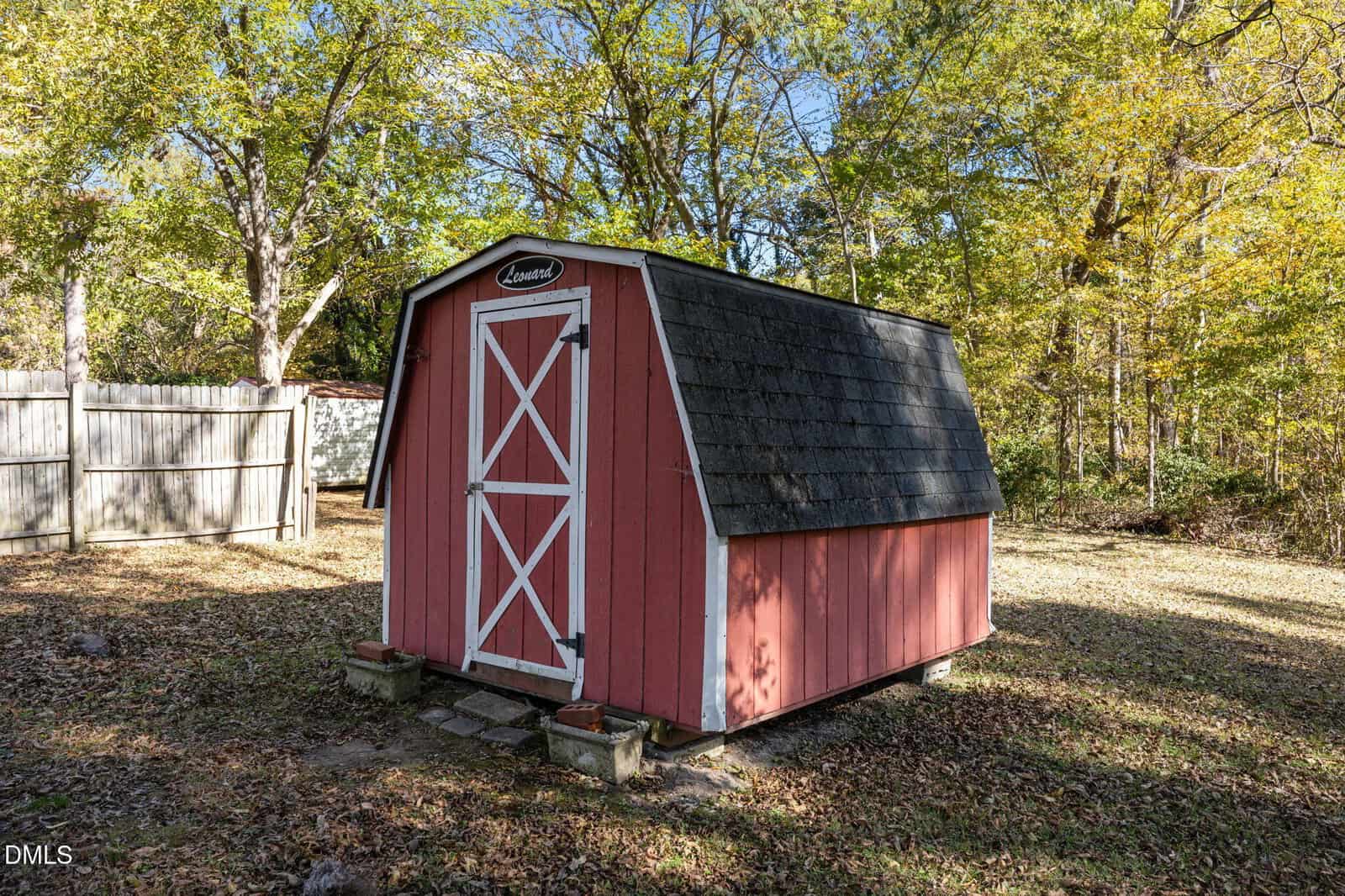 A small red wooden shed with white trim and double doors sits on a grassy yard surrounded by trees and a wooden fence.
