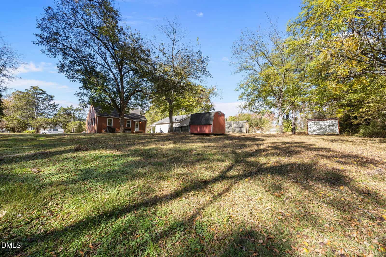 A grassy yard with scattered trees, a red house to the left, a red shed and a white building in the center, and another small white shed to the right, all under a blue sky.