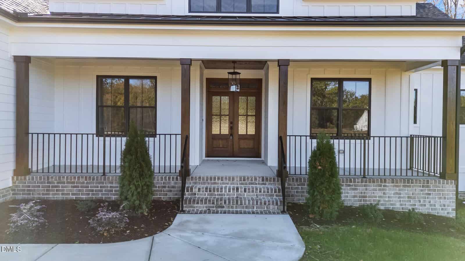 A front view of a house with a covered porch, double wooden doors, black framed windows, brick steps, metal railings, and landscaped shrubs in front of the porch.