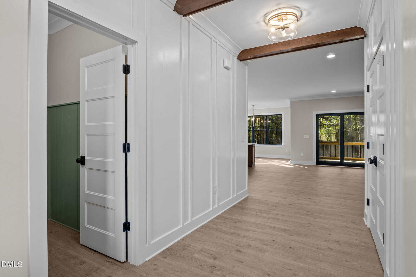 A hallway with light wood flooring, white paneled walls, and visible wooden ceiling beams leads to a living area with large windows and glass doors that open to a deck. A small green accent wall is visible in an adjoining room.