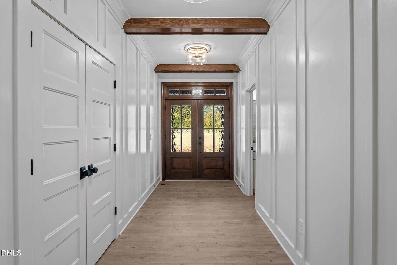 A hallway with light wood flooring, white paneled walls, and wooden ceiling beams leads to double front doors with glass panels. There are multiple white doors with black handles on the left wall.
