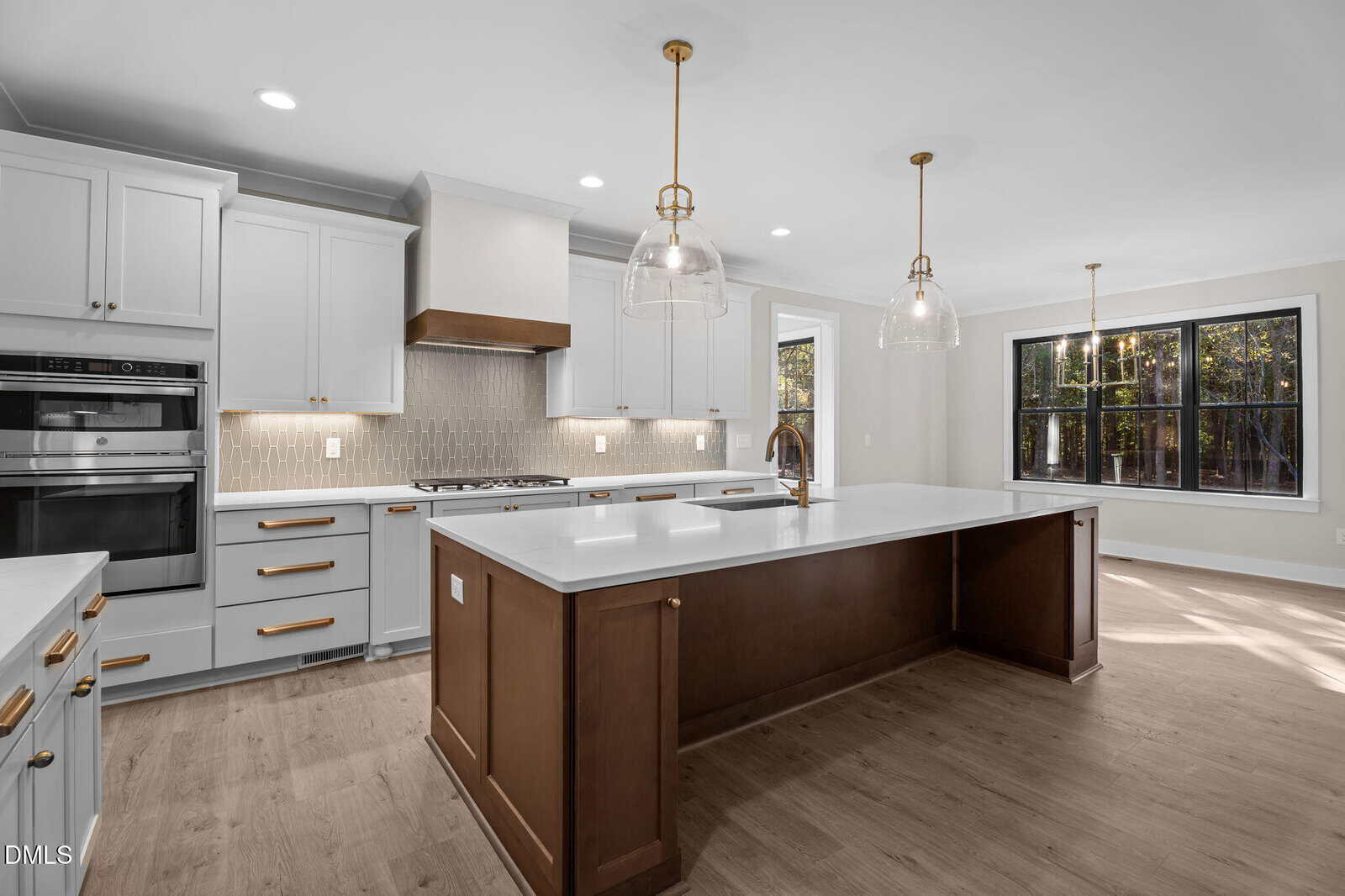 Modern kitchen with white cabinets, wood accents, double wall ovens, gas cooktop, large island with sink, pendant lights, light wood floors, and a dining area with large windows showing trees outside.