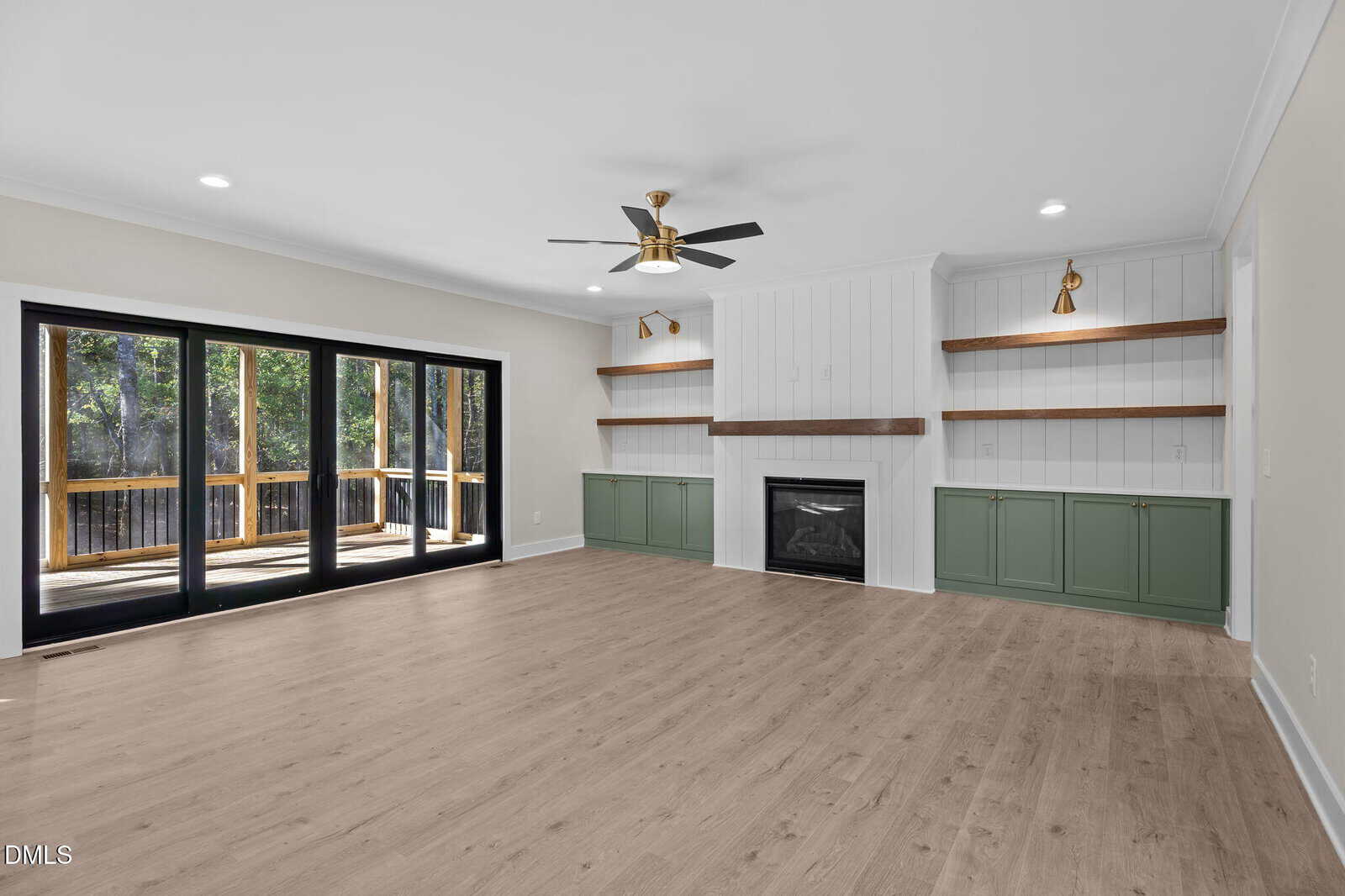 A spacious living room with light wood flooring, a ceiling fan, built-in shelves with green cabinets, a fireplace, and large sliding glass doors opening to a wooden deck with a view of trees.