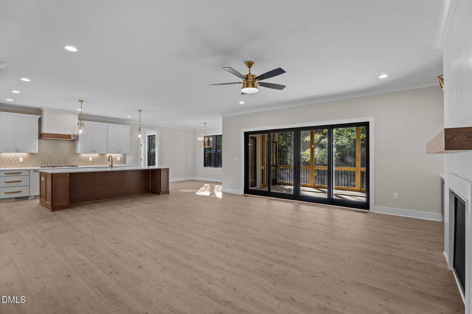 Open-concept living space with light wood flooring, white walls, a modern ceiling fan, a fireplace, and large sliding glass doors leading to a deck. The kitchen has white cabinets, an island, and pendant lights.