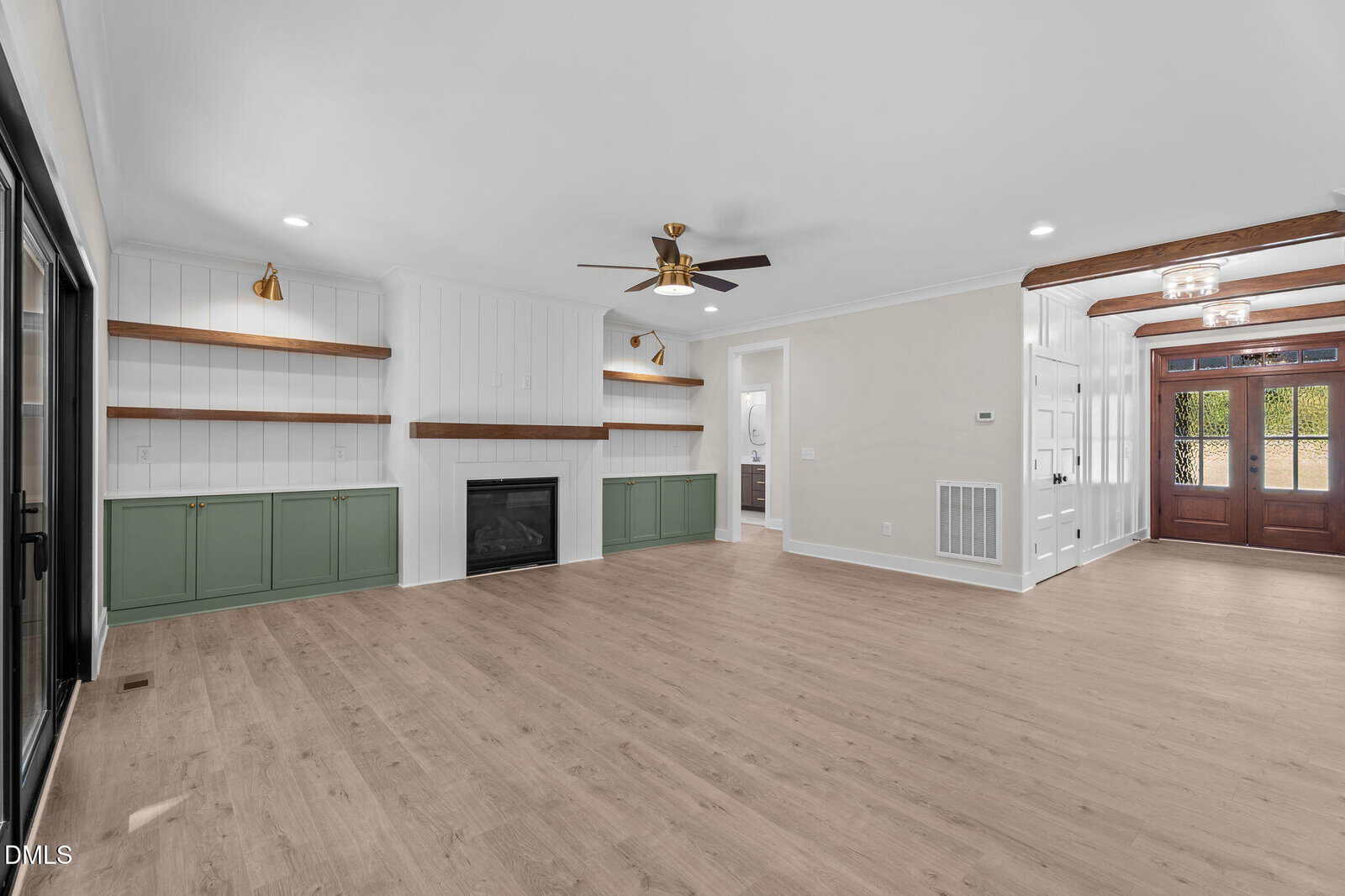 A spacious living room with light wood flooring, white walls, a fireplace flanked by green cabinets with wooden shelves, ceiling fan, exposed beams, and a front entry with glass double doors.