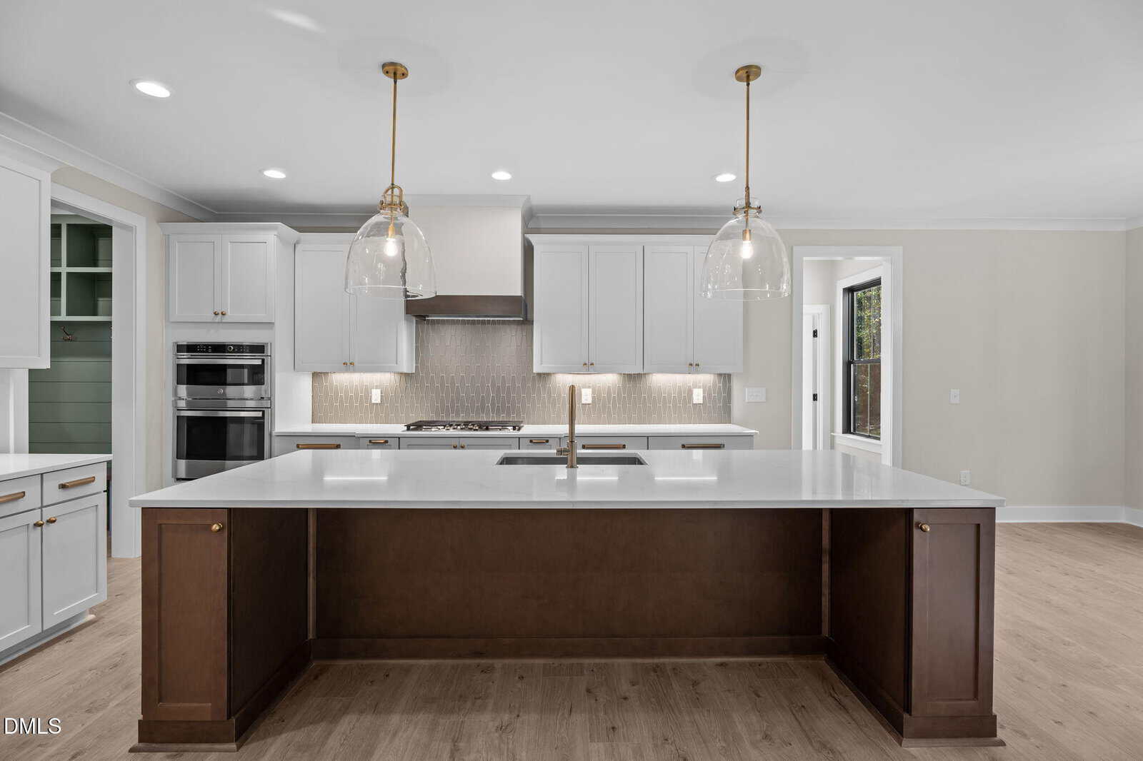 Modern kitchen with white cabinets, stainless steel appliances, a large island with a brown base, two pendant lights above, and light wood flooring. Natural light enters from a door and windows on the right.