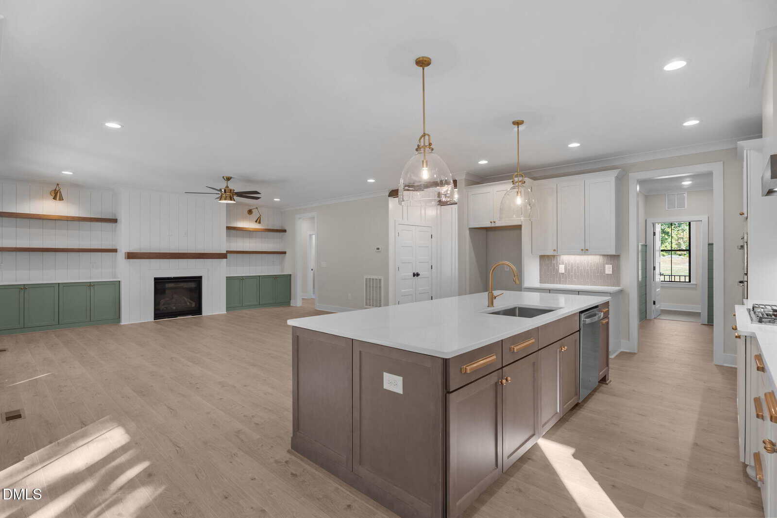 Modern open-concept kitchen and living area with light wood floors, white cabinetry, large island, gold fixtures, built-in shelves, fireplace, and recessed lighting. A hallway and doorways are visible in the background.