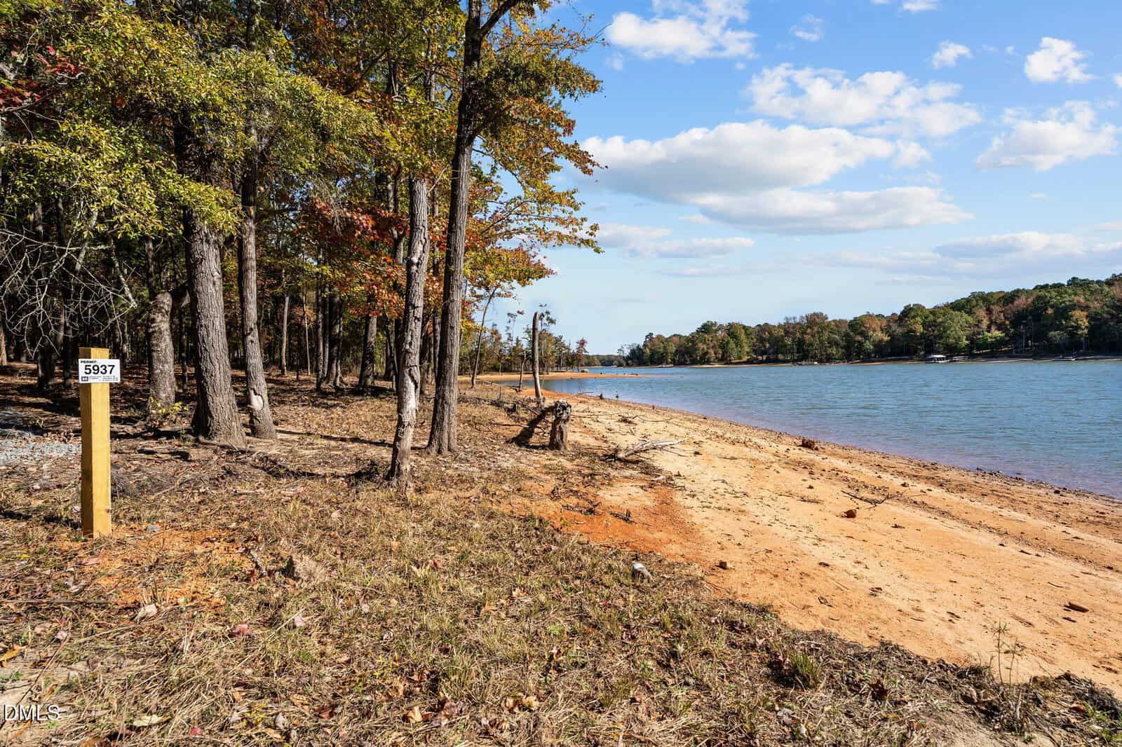 A sandy shoreline curves beside a body of water. Trees with autumn foliage line the left side, and a wooden post marked "5937" stands near the shore. The sky is mostly clear with a few clouds.