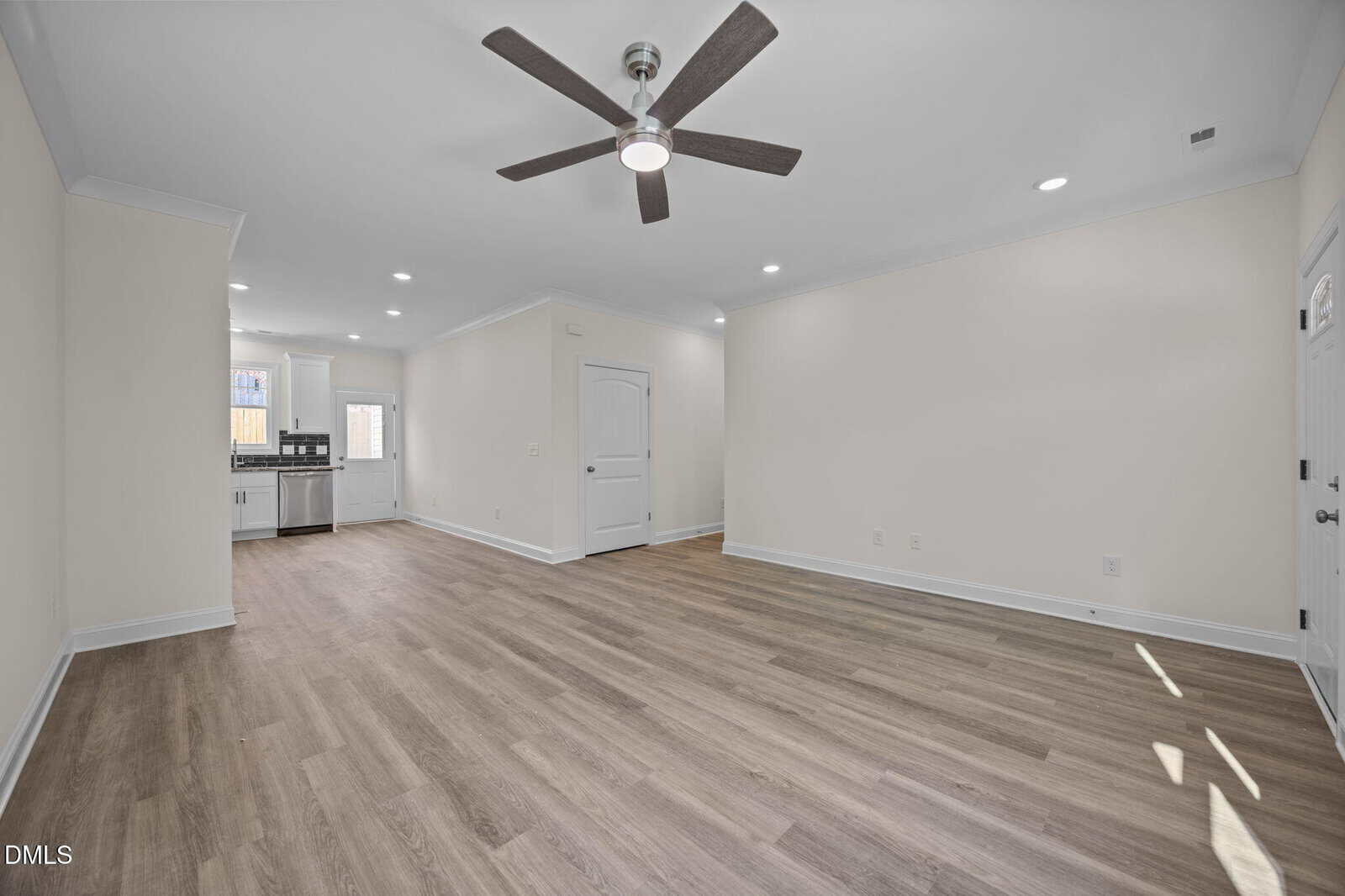 Open living area with light wood flooring, white walls, ceiling fan, and recessed lighting. Kitchen with stainless steel appliances and white cabinetry is visible in the background. Multiple doors and natural light from windows.
