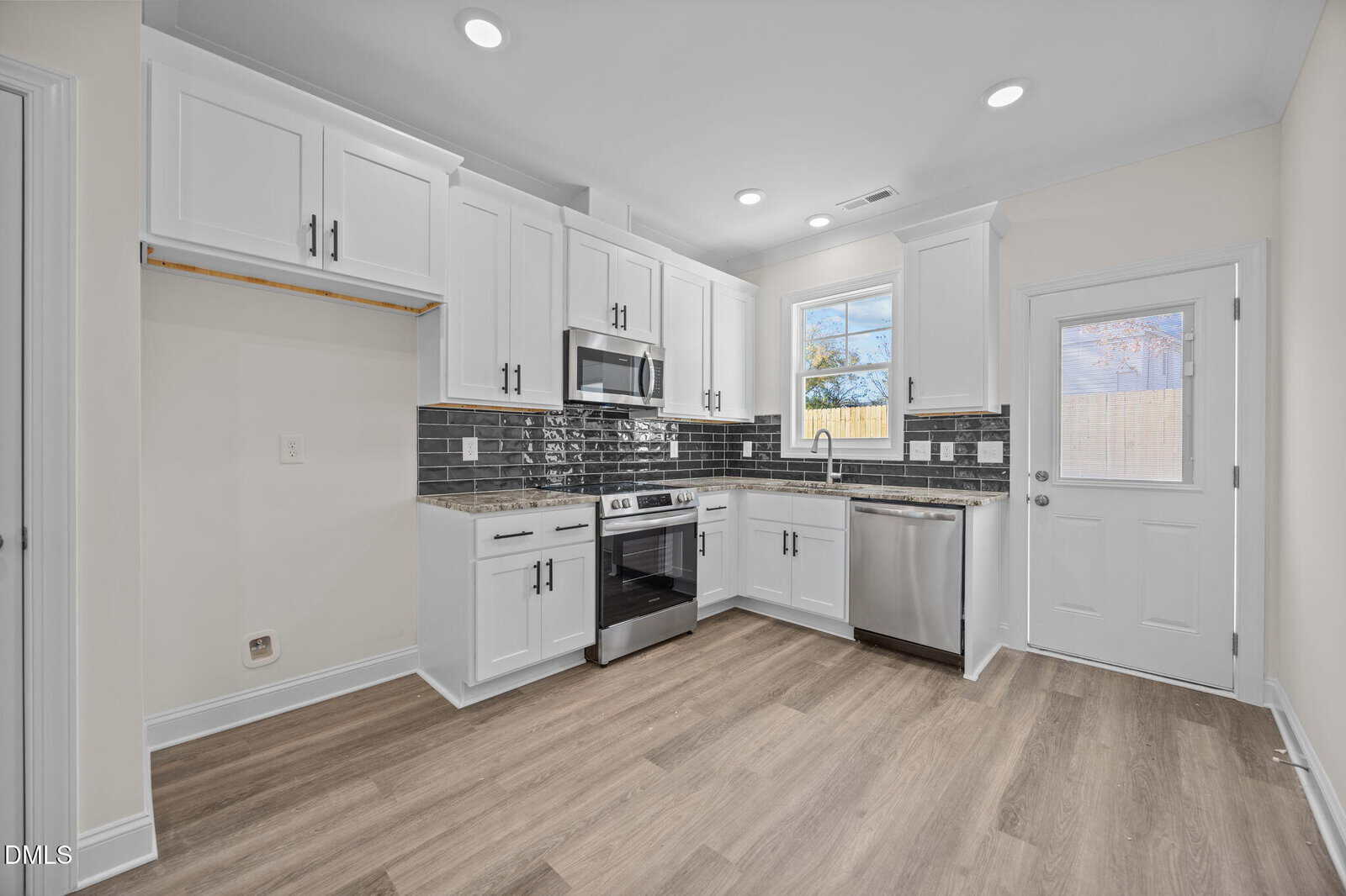 Modern kitchen with white cabinets, stainless steel appliances, black tile backsplash, a window above the sink, light wood flooring, and a white door leading outside. The space is brightly lit with ceiling lights.