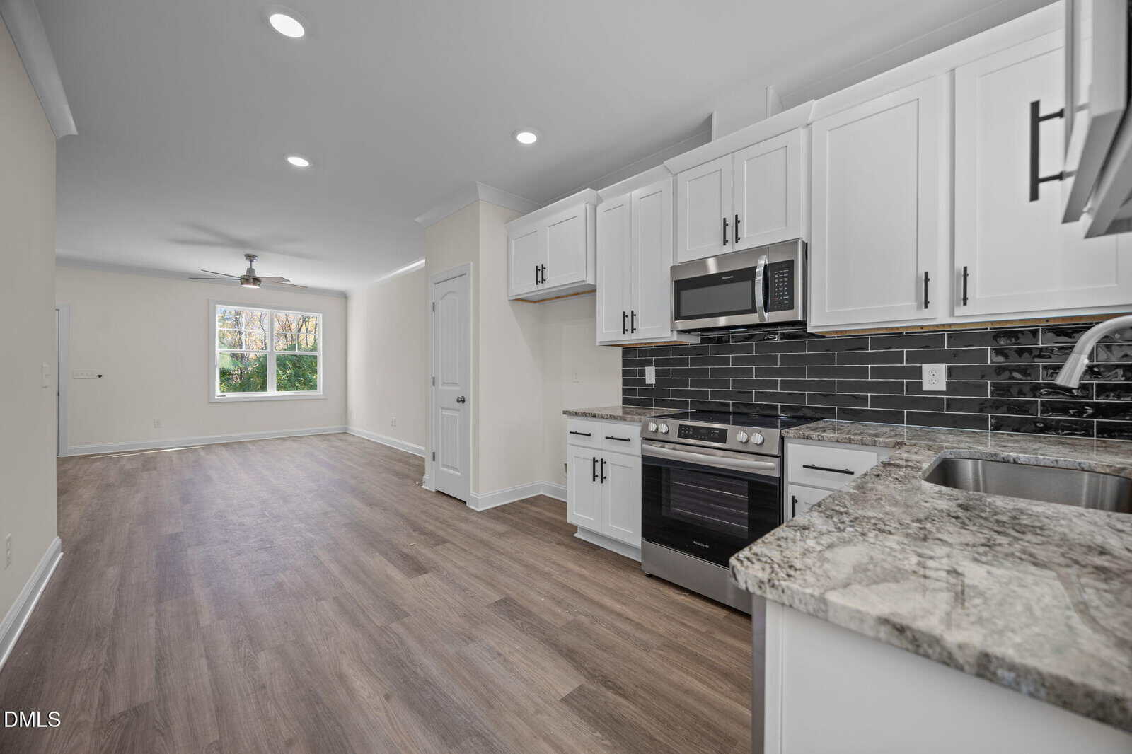 Modern kitchen with white cabinets, stainless steel appliances, black tile backsplash, and marble countertops, opening into a spacious living area with wood flooring and a large window letting in natural light.