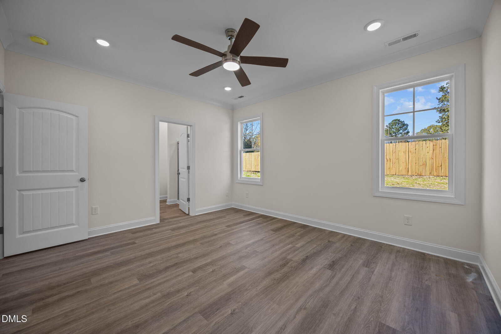 Bright, empty room with wood flooring, ceiling fan, and recessed lights. Two windows show a fenced backyard. Doorways lead to another room and a closet or bathroom. Walls are light-colored.