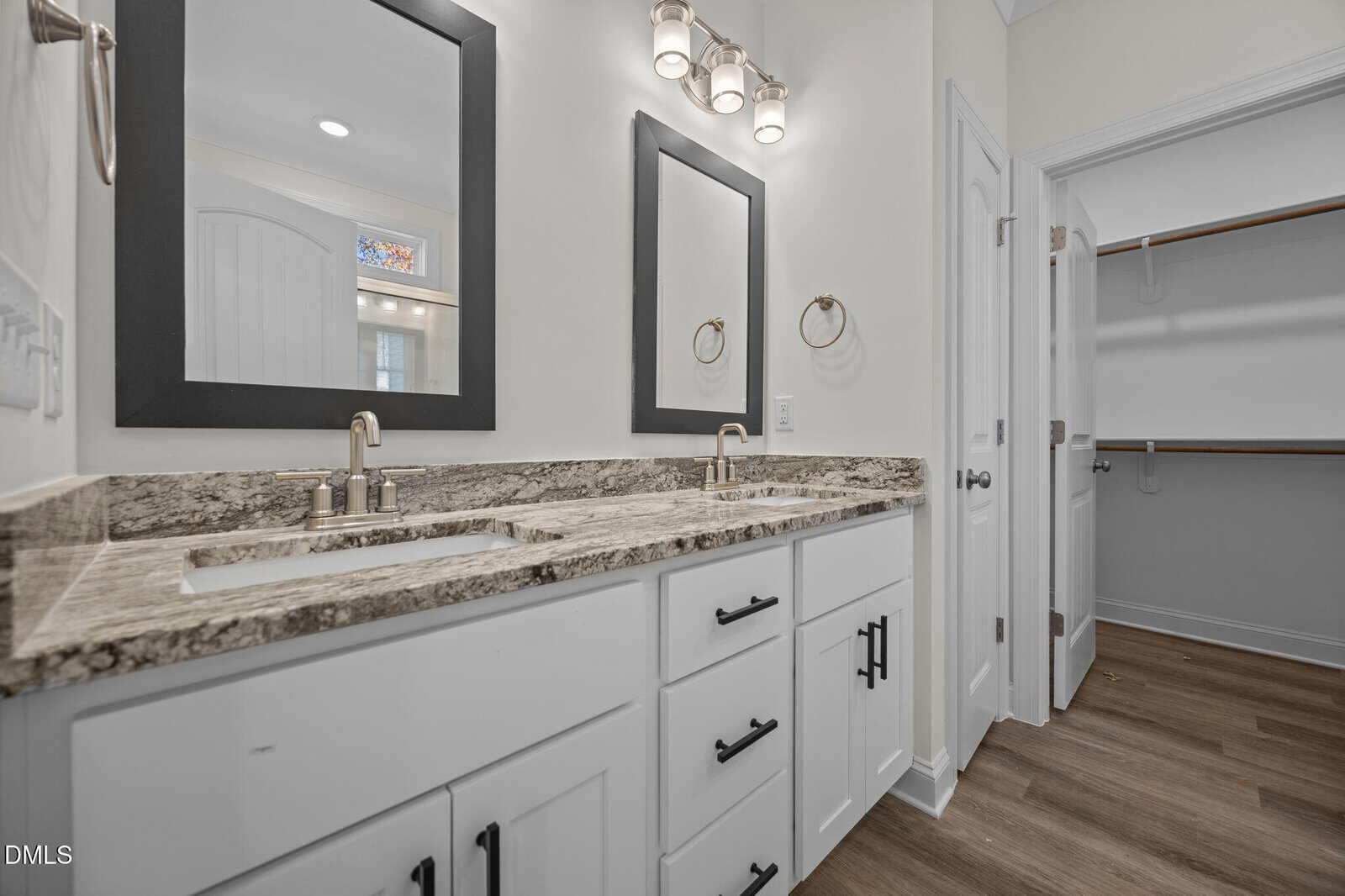 Two rectangular mirrors and double sinks with granite countertop in a modern bathroom with white cabinets, black hardware, and wood-style flooring. An open closet with shelves is visible through the door on the right.
