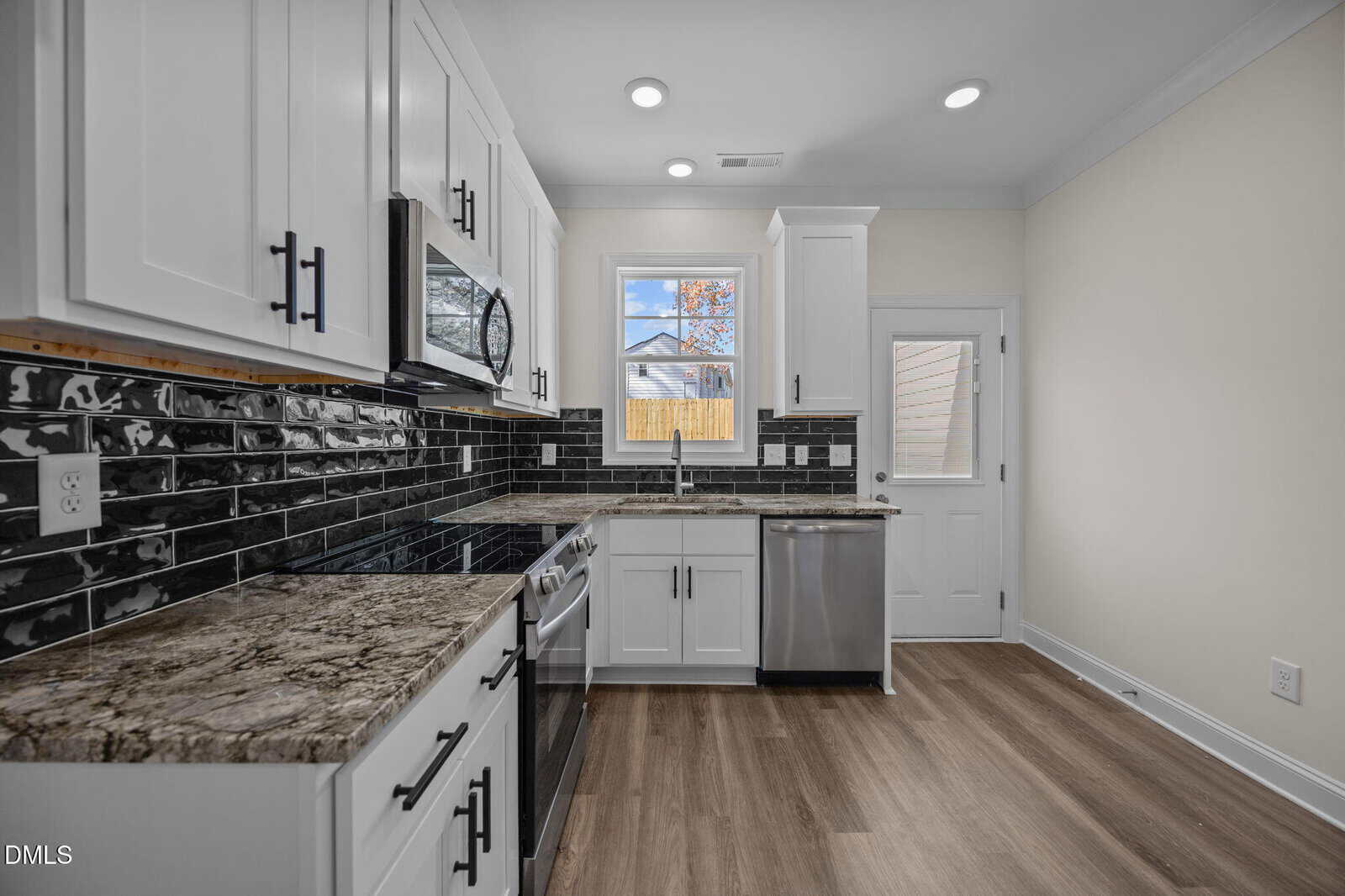Modern kitchen with white cabinets, black tile backsplash, stainless steel appliances, marble countertops, wood flooring, a window above the sink, and a door leading outside.