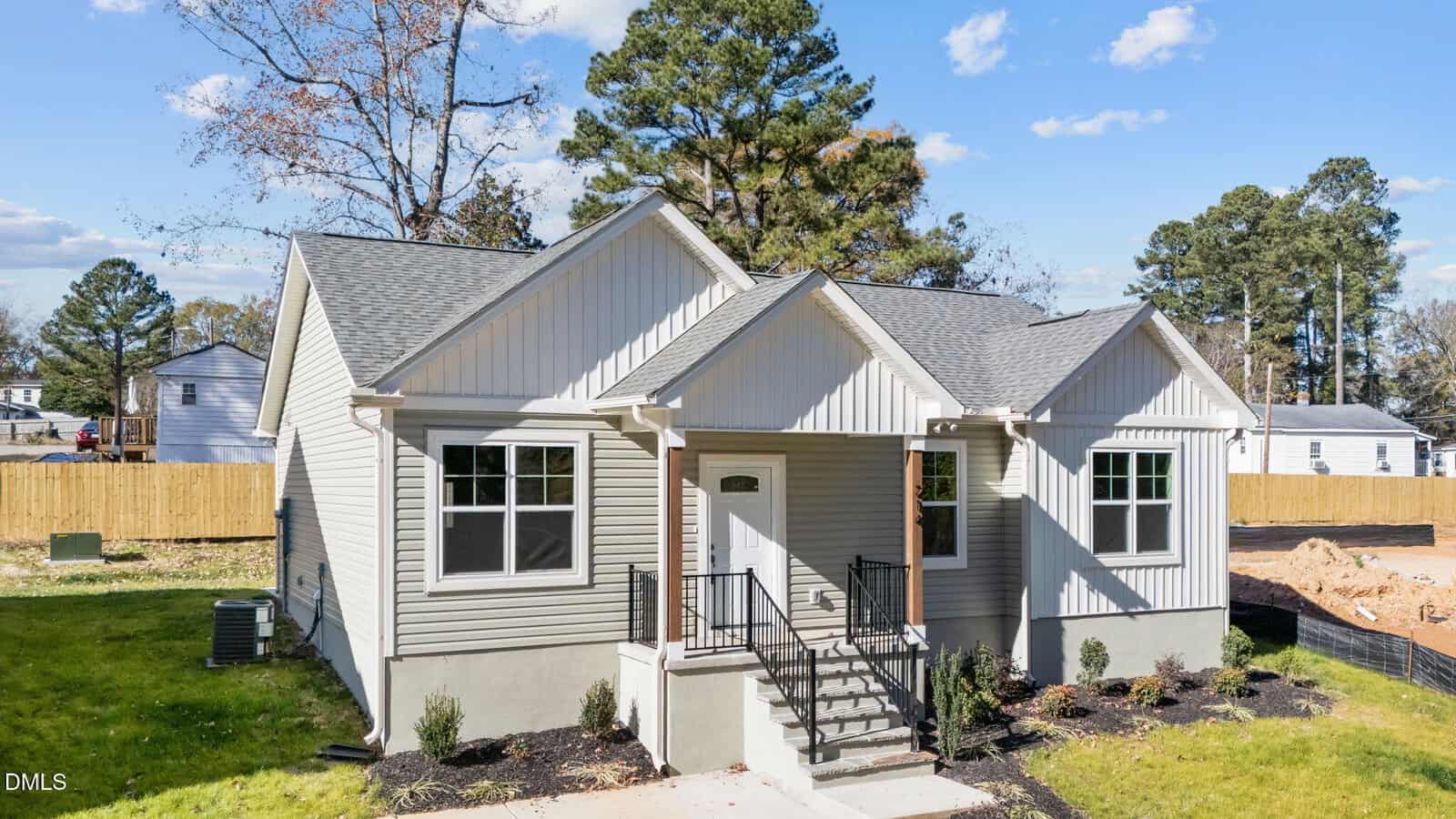 A modern, single-story house with light gray siding, white trim, and a small porch with black railings. The yard has new landscaping and is surrounded by a wooden fence, with trees and blue sky in the background.