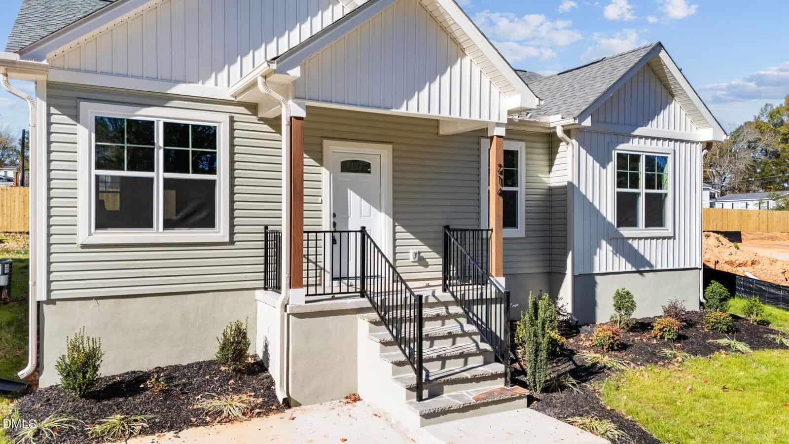 A small, newly built house with light-colored siding, white trim, and a covered front porch. Steps with a black metal railing lead to the front door. Fresh landscaping and green grass surround the home.
