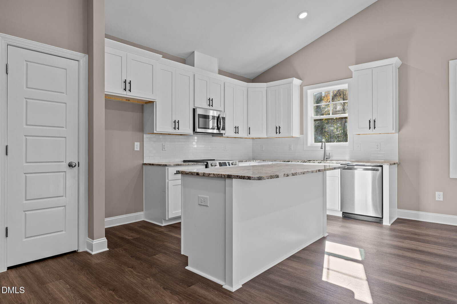 Bright modern kitchen with white cabinets, stainless steel appliances, a center island, marble countertops, and wood flooring. Sunlight streams in from a window above the sink, highlighting the open, clean space.