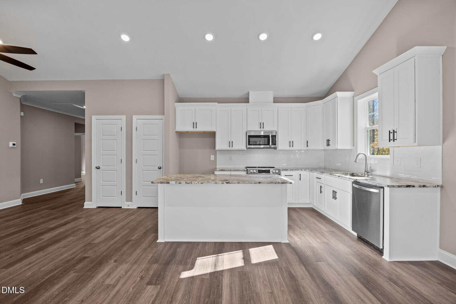Modern kitchen with white cabinets, stainless steel appliances, marble countertops, and wood flooring. The space is illuminated by recessed lighting and natural light from a window above the sink.