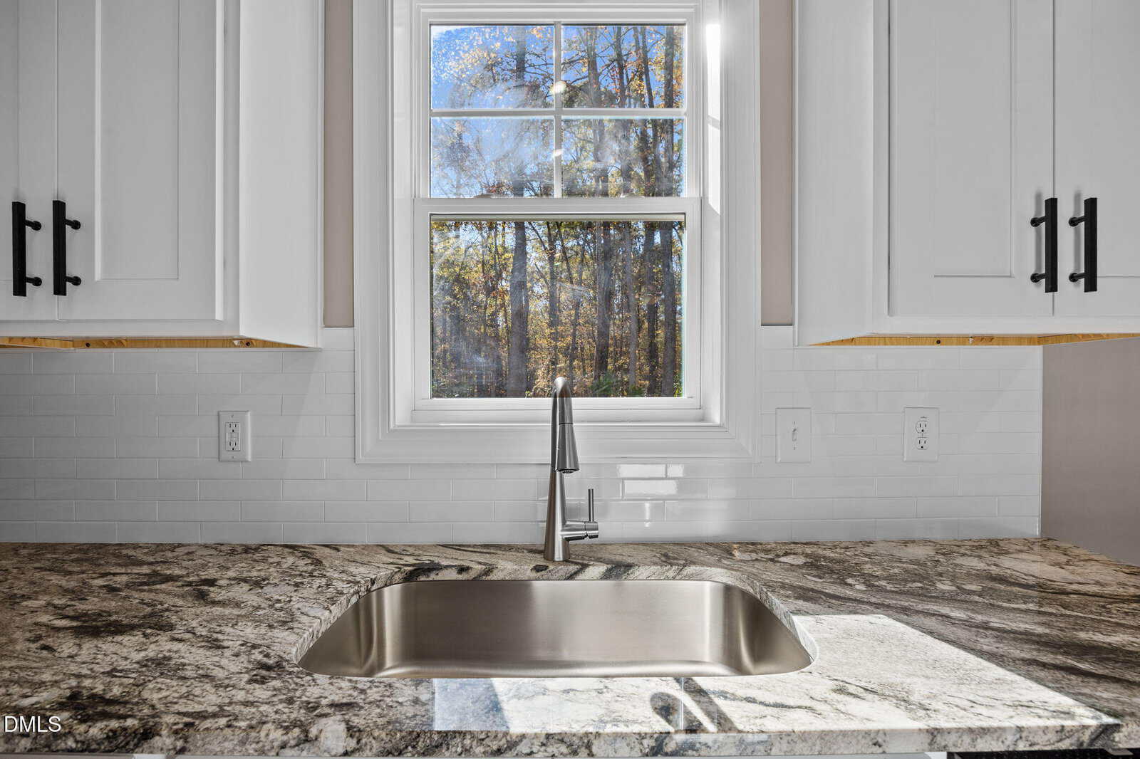 A modern kitchen sink with a stainless steel faucet sits beneath a window, surrounded by white cabinets and a marble countertop. Sunlight streams through the window, revealing trees outside.