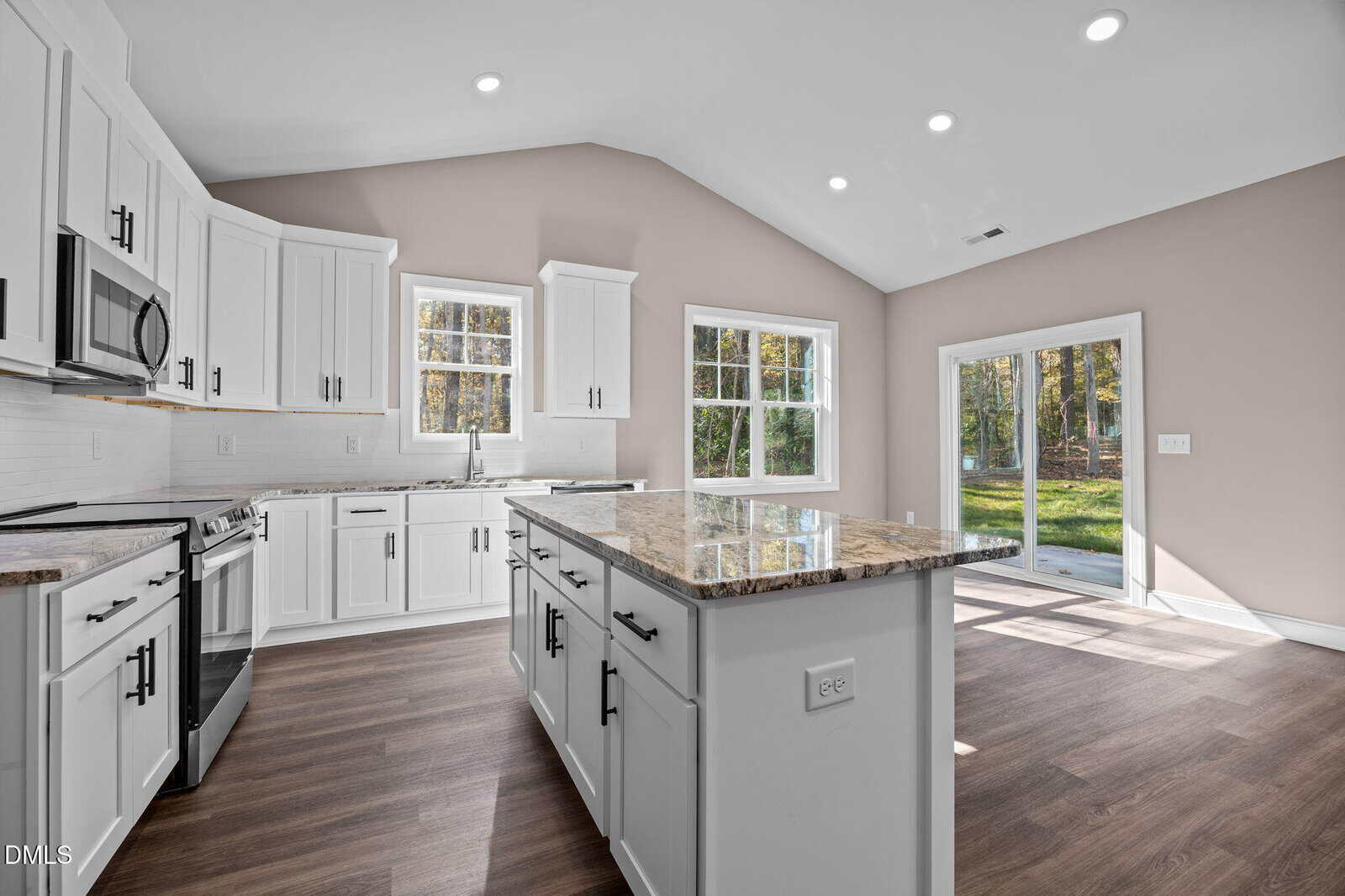 Modern kitchen with white cabinets, stainless steel appliances, a center island with granite countertop, wood flooring, large windows, and glass doors leading to a green backyard. Natural light fills the space.