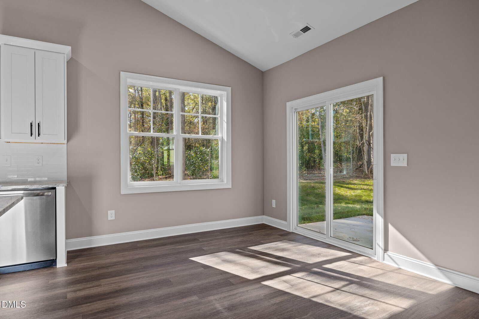 Sunlit room with light brown walls, dark wood floors, white trim, a window, and glass sliding doors opening to a grassy backyard. Part of a stainless steel dishwasher and white cabinet are visible on the left.