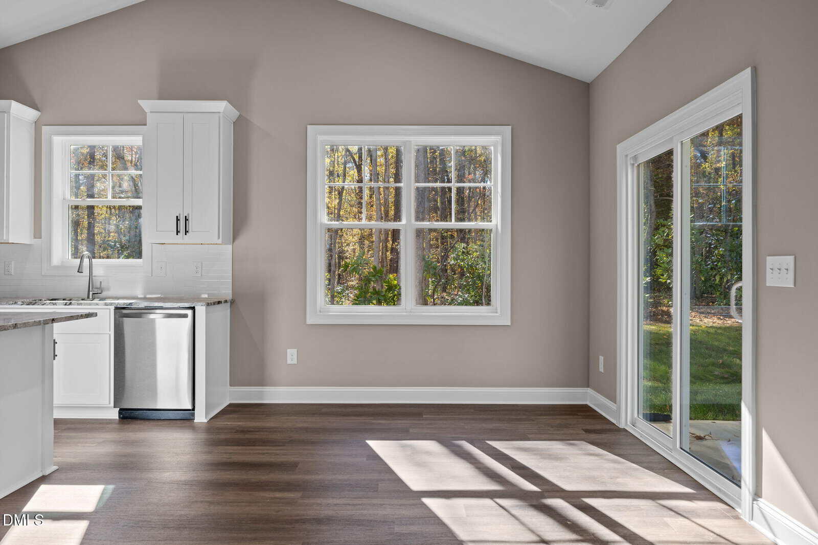 Bright, modern kitchen and dining area with large windows, sliding glass door to a green yard, white cabinets, stainless steel appliances, and wood flooring. Sunlight streams onto the floor.