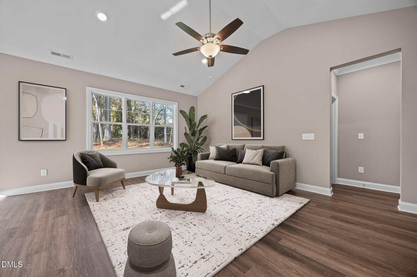 Modern living room with a gray sofa, armchair, large green plant, coffee table, patterned rug, and framed art. Large windows let in natural light, and ceiling fan is on. The walls are painted light beige.