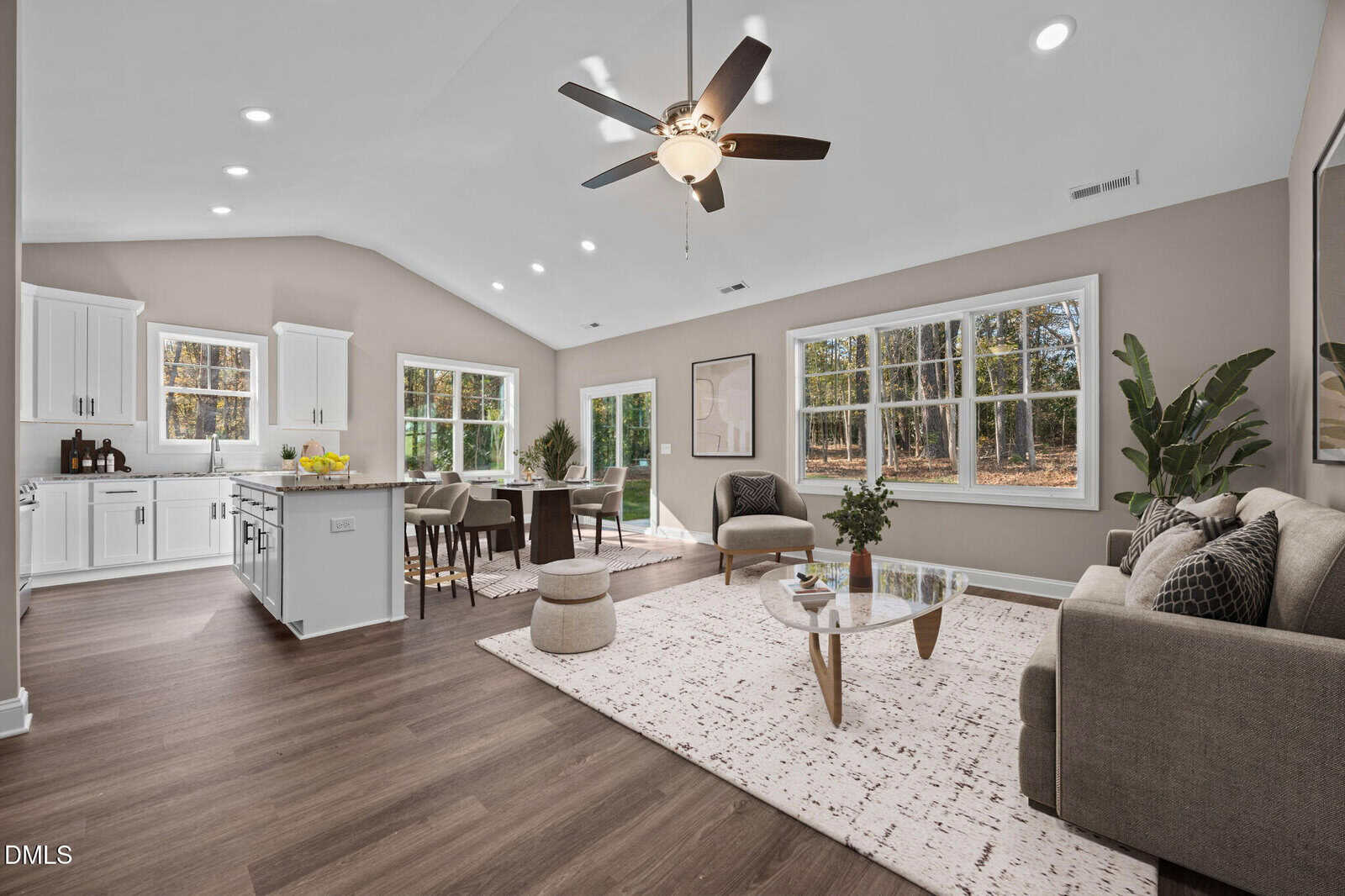 Spacious open-concept living room and kitchen with large windows, modern furnishings, wood floors, neutral walls, and abundant natural light, overlooking a wooded backyard.