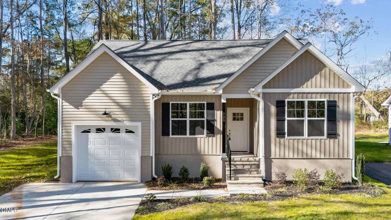 A small, beige single-story house with white trim, black shutters, and an attached single-car garage sits at 220 E College Street in Franklinton. Front steps with a railing lead to a neat lawn surrounded by trees under a blue sky.