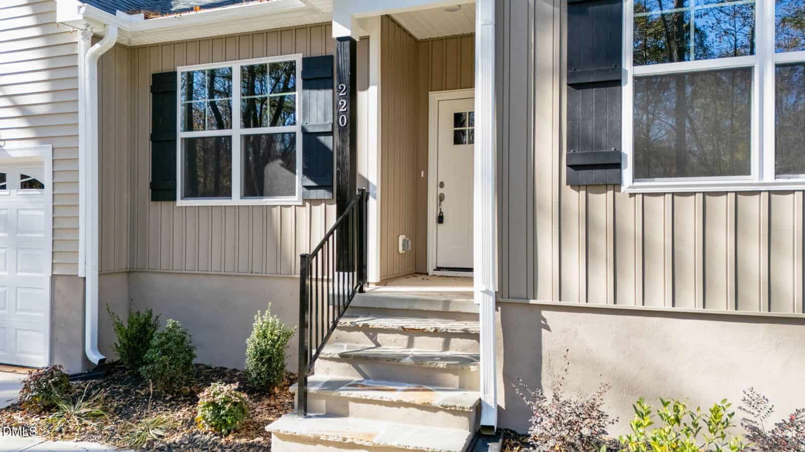 Front entrance of a beige house with black shutters, a white door with a window, black metal railing on stone steps, and small shrubs along the walkway.