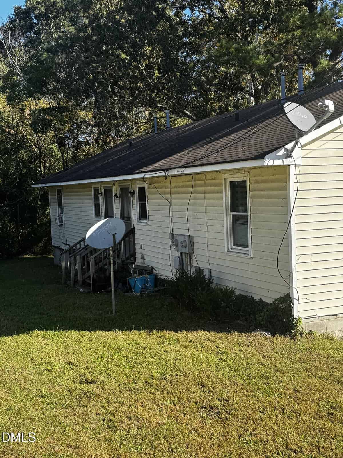 A one-story house with white siding, several satellite dishes attached, a small wooden staircase, visible utility wires, and a grassy yard; trees are in the background.
