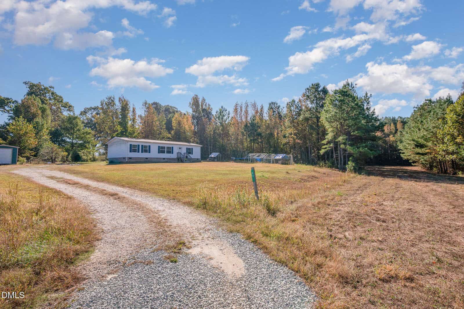 A gravel driveway curves toward a single-story white manufactured home with green shutters at 93 Grove Park Lane, Henderson, set on a grassy lot bordered by trees. Several small greenhouse structures are visible behind the house.
