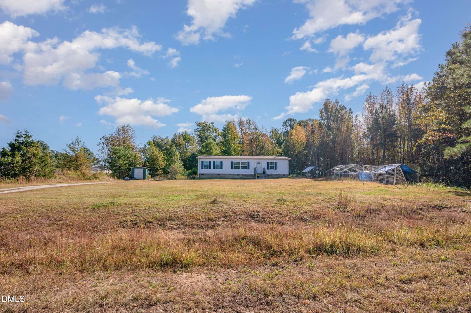 A single-story white house sits in the center of a large, grassy yard with trees in the background, a small shed to the left, and two metal structures to the right under a blue sky with scattered clouds.